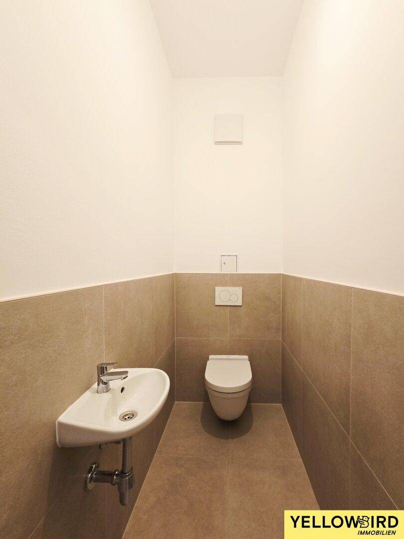 Separates WC mit modernem Wand-WC und kleinem Waschbecken, in neutralen Beigetönen gefliest.