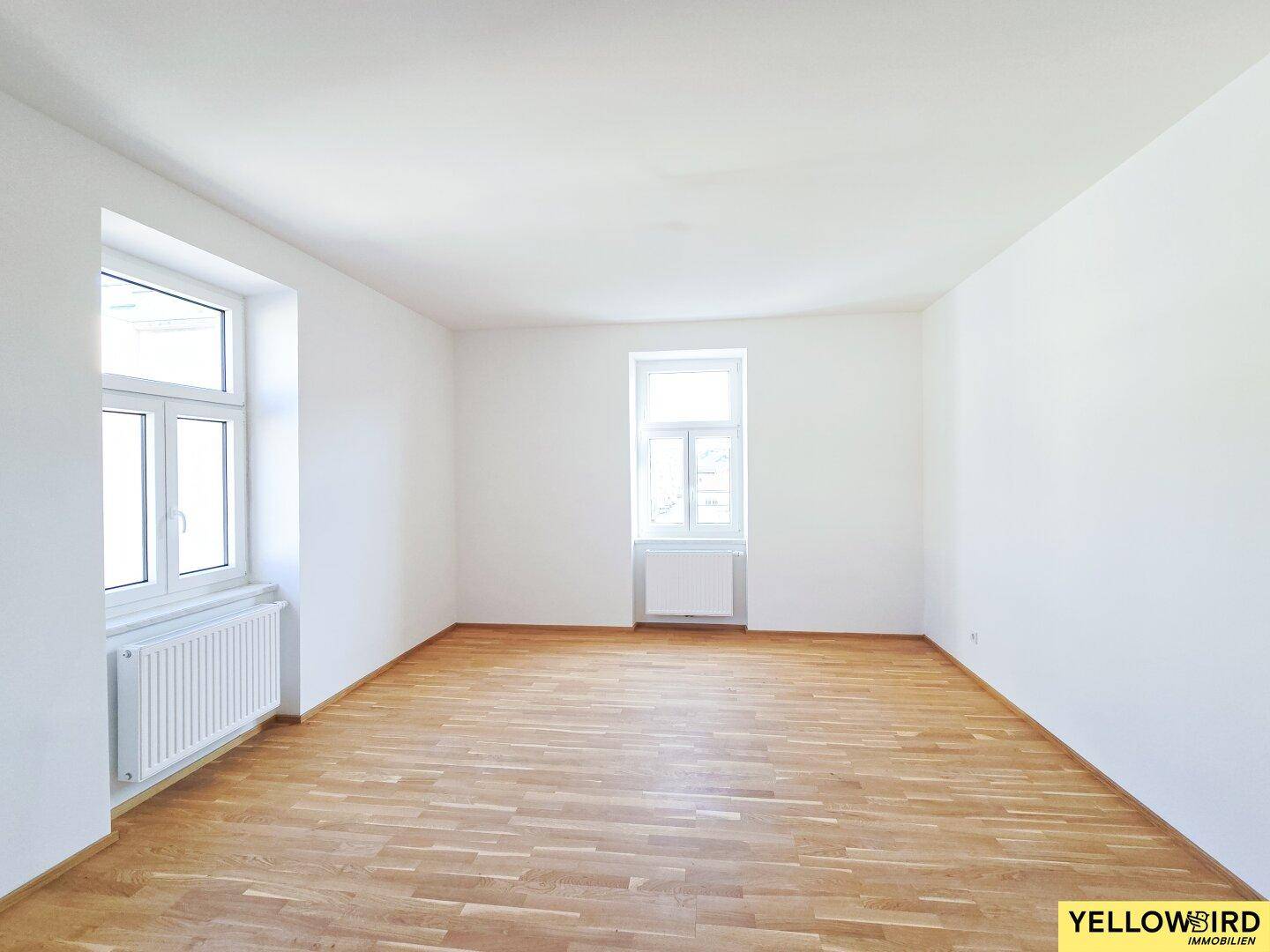 Geräumiges, leeres Zimmer mit hellem Parkettboden und zwei Fenstern, bereit zur individuellen Gestaltung.