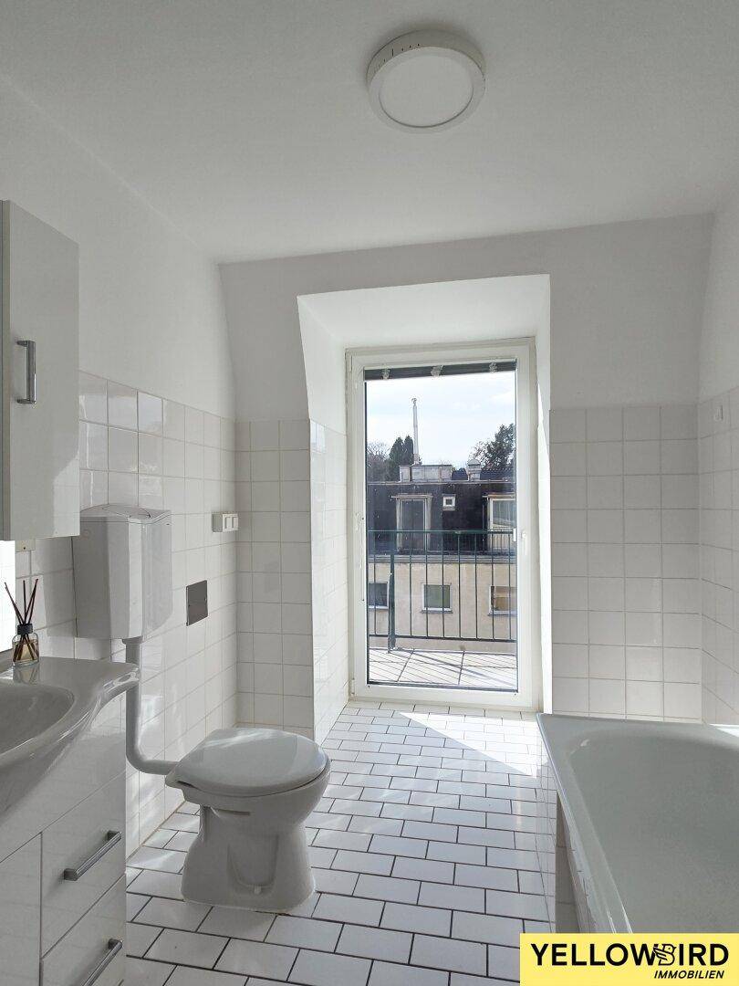 Helles Badezimmer mit weißen Fliesen, Badewanne und Fenster mit Ausblick.