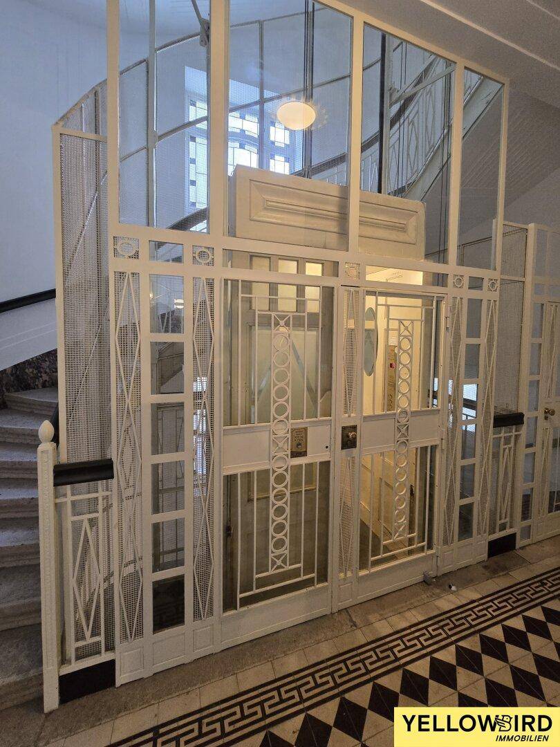 Historischer Aufzug in einem eleganten Treppenhaus mit kunstvollen Gittern und Fliesenboden.