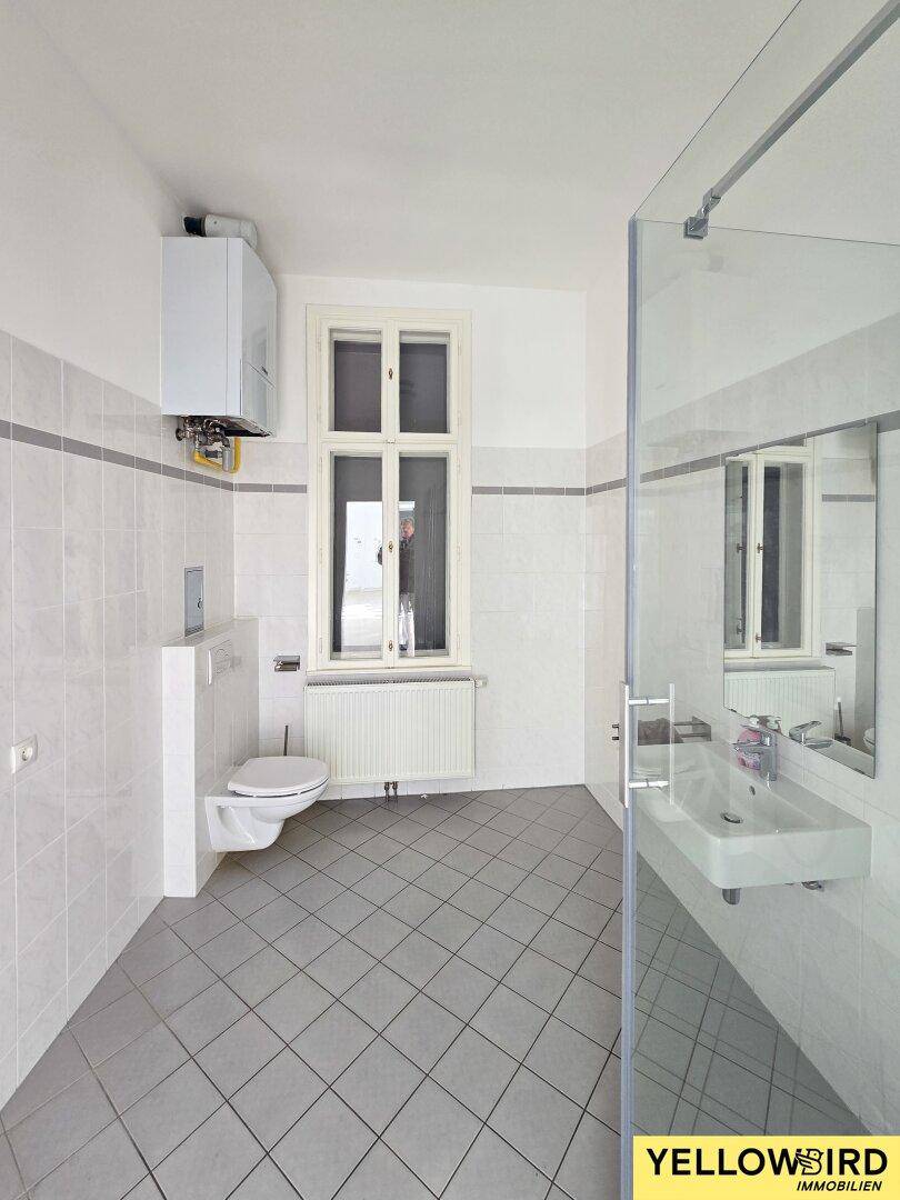 Helles Badezimmer mit Dusche, WC und Fenster, ausgestattet mit grauen Bodenfliesen.