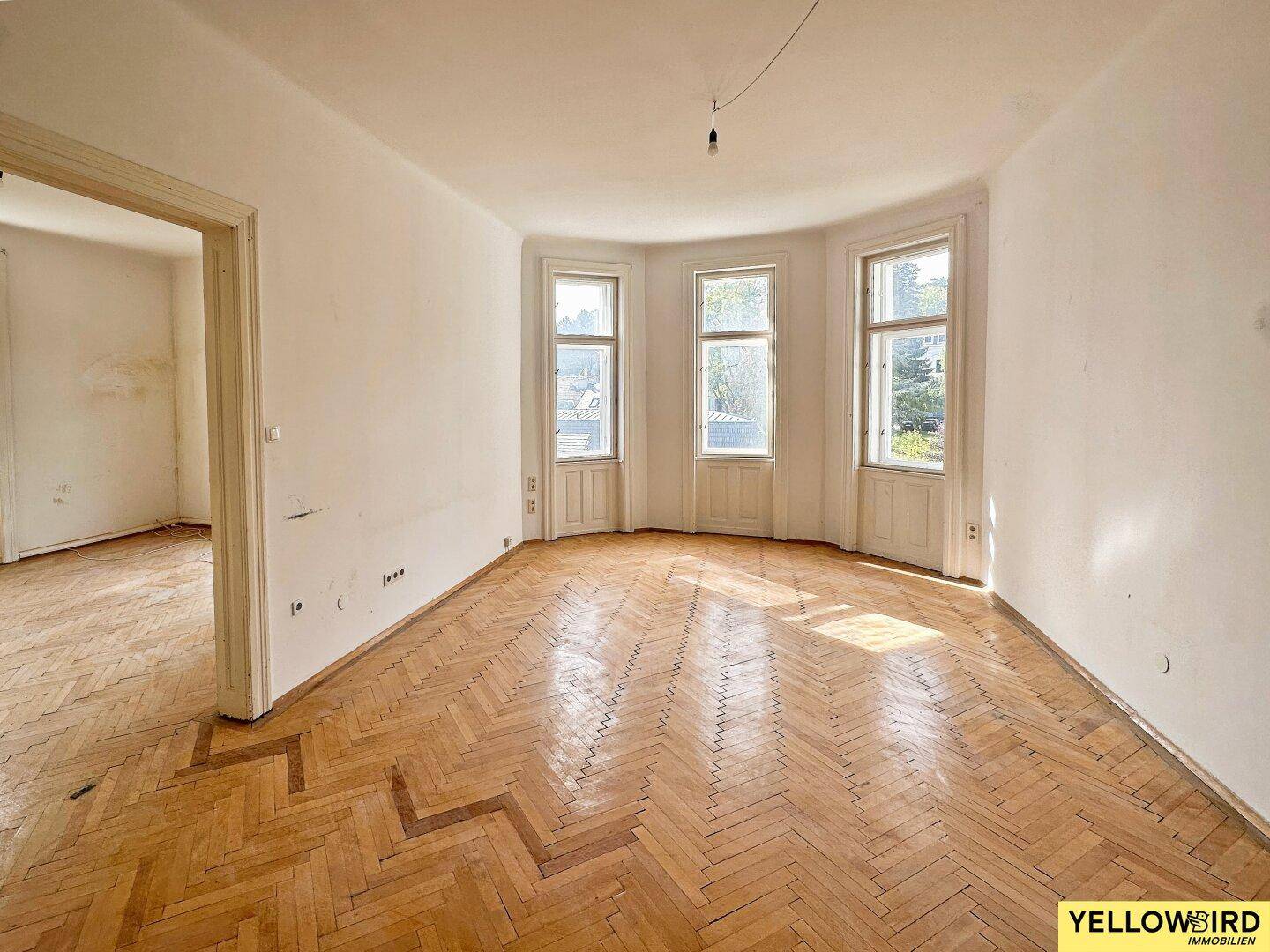 Leerer Raum mit Fischgrätparkett und Erkerfenstern, bereit für Renovierung.