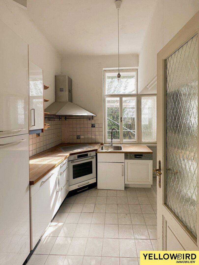 Einfache Küche mit weißen Schränken, Holztheke und Fliesenboden, renovierungsbedürftig.