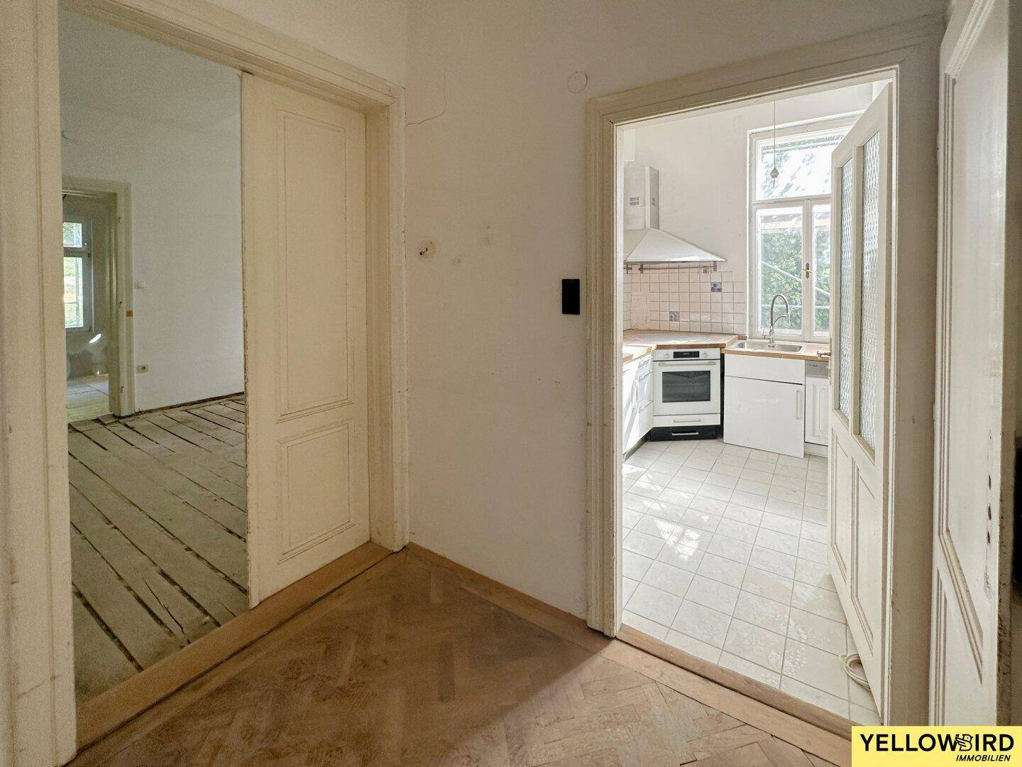 Flur mit Fischgrätparkett und Blick in eine einfache Küche mit Fliesenboden.