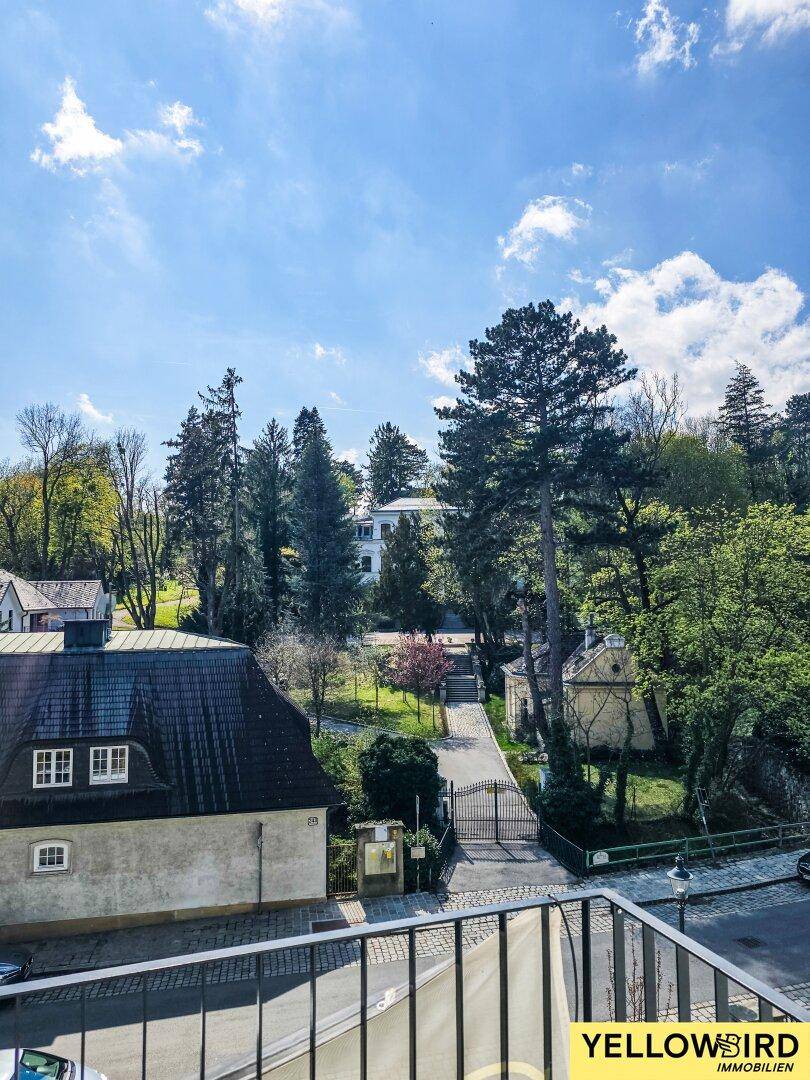 Blick vom Balkon auf die grüne Umgebung mit Straße, Tor und benachbarten Gebäuden.