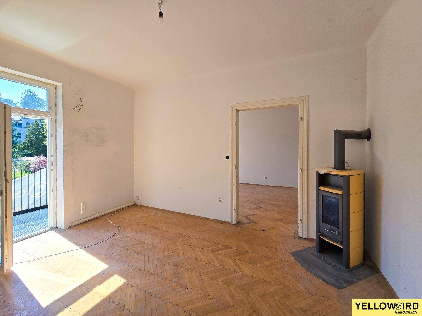 Leerer Raum mit Fischgrätparkett, Holzofen und Balkonzugang, ideal für eine Renovierung.