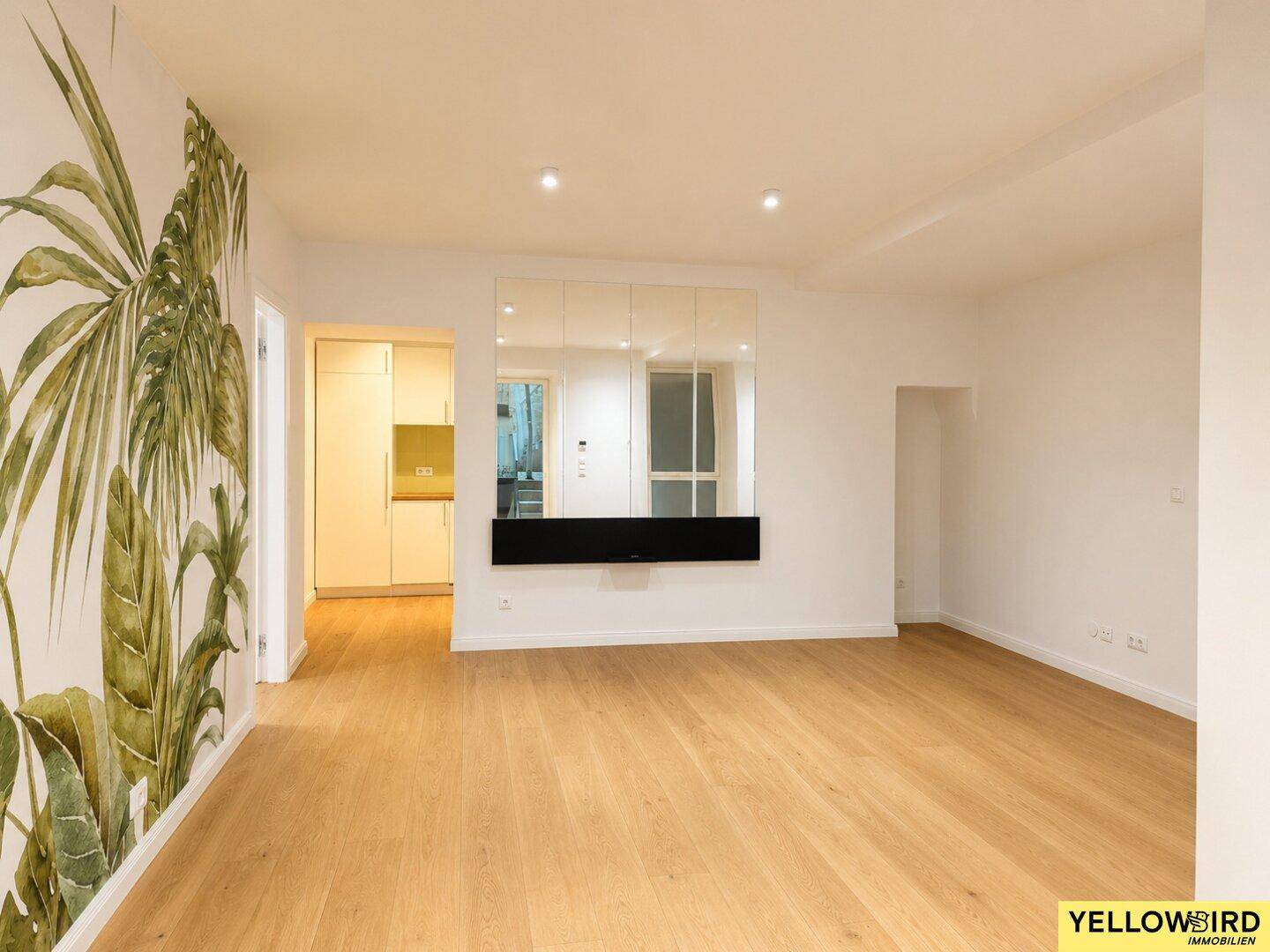 Wohnzimmer mit Holzboden, tropischer Wandtapete und Blick auf eine angrenzende Küchenzeile.
