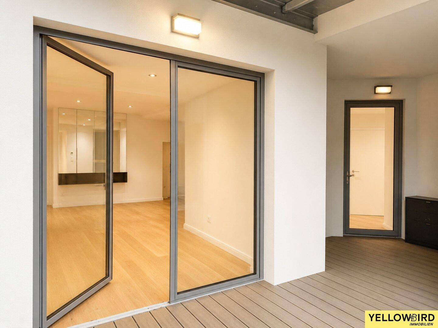 Zugang zum Gebäude über eine Holzterrasse mit großen Glastüren und Außenbeleuchtung.