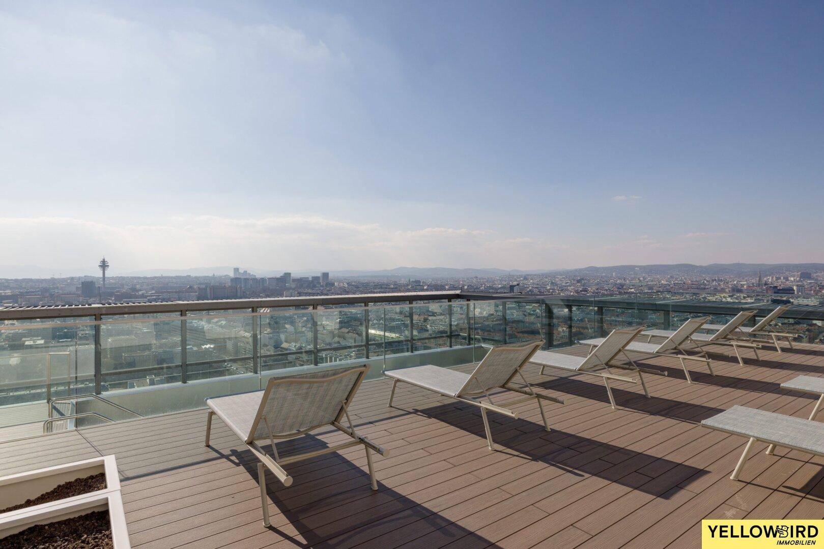 Entspannungsbereich auf der Dachterrasse mit Liegestühlen und beeindruckendem Stadtpanorama unter blauem Himmel.