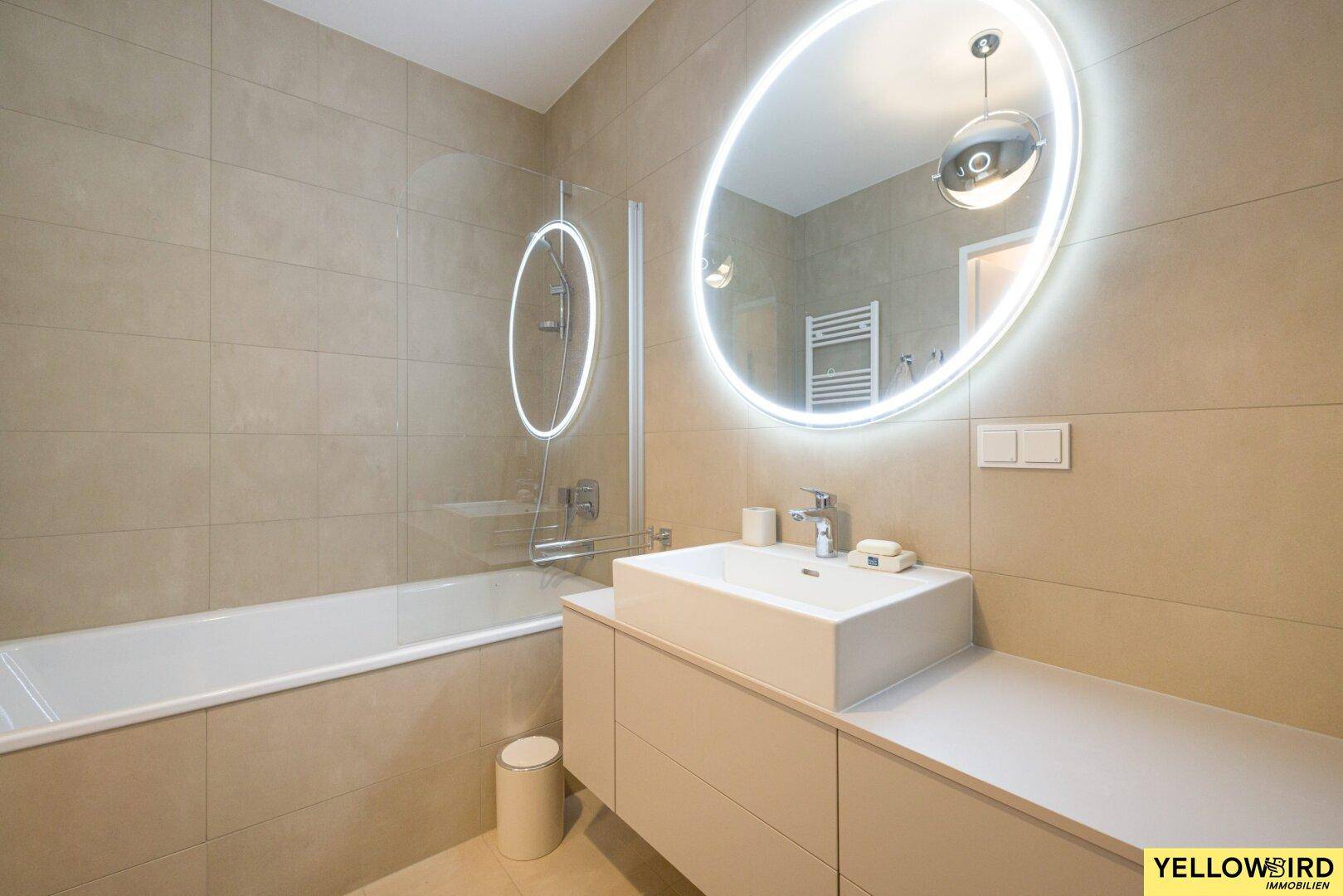 Modernes Badezimmer mit beigefarbenen Fliesen, Badewanne und beleuchtetem Spiegel.