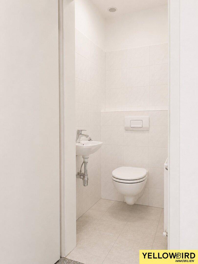 Separates WC mit wandhängender Toilette, kleinem Waschbecken und hellen Fliesen.