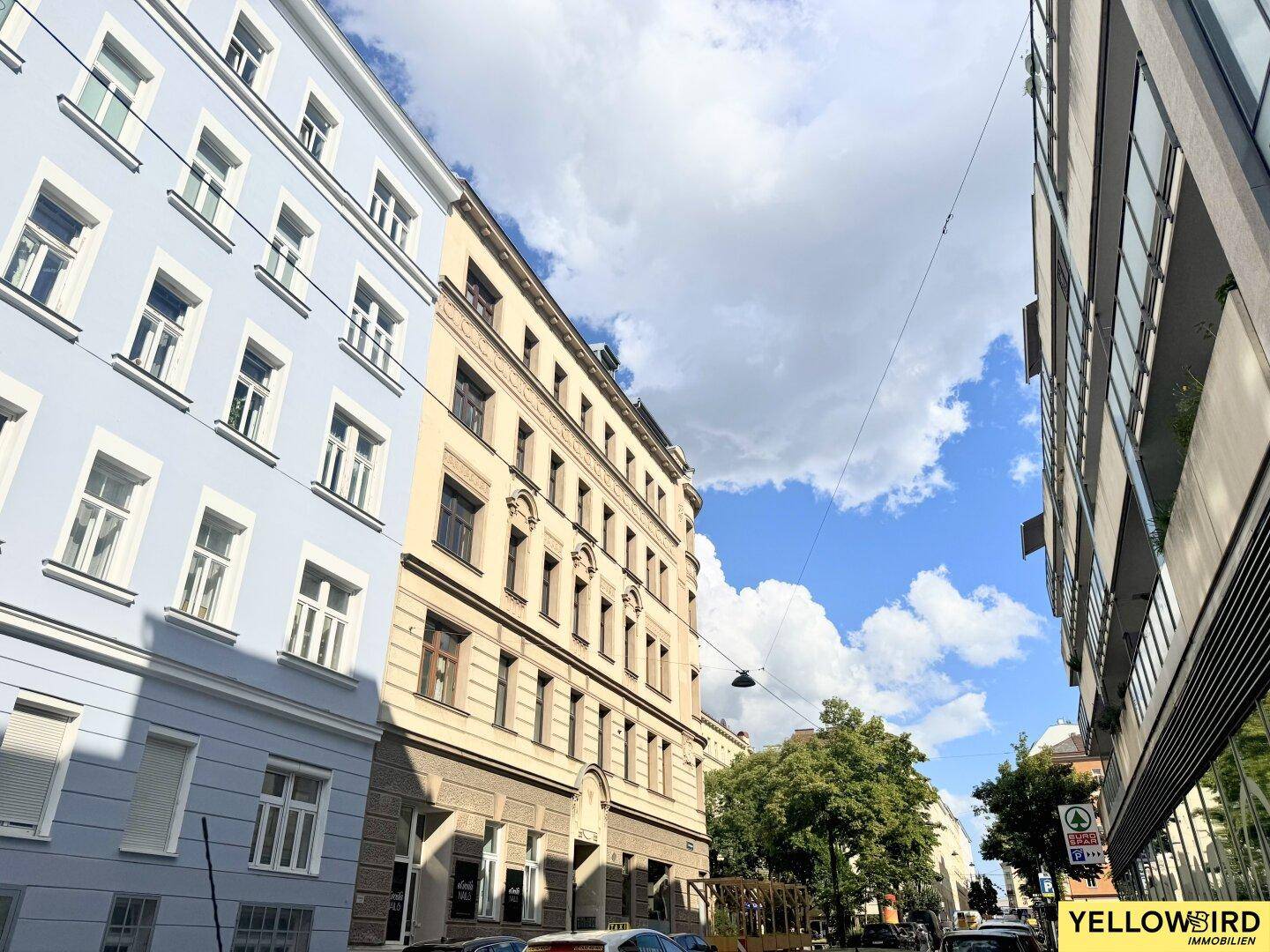 Straßenansicht mit historischen Gebäuden und Bäumen unter blauem Himmel.