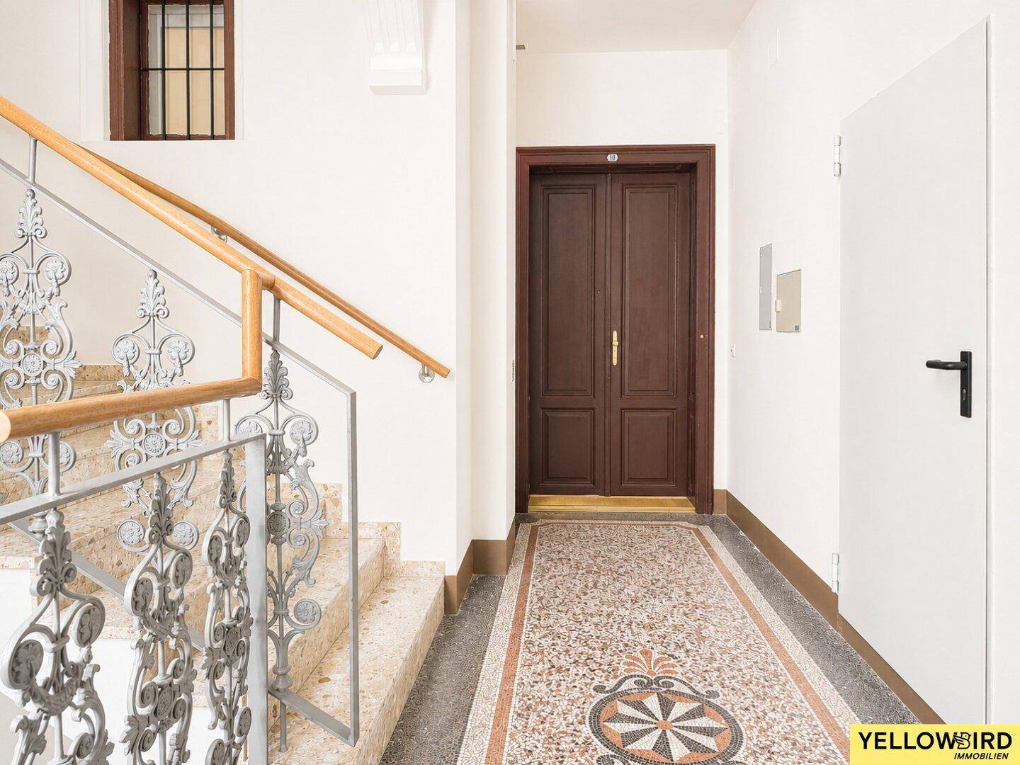 Historisches Treppenhaus mit kunstvollem Geländer, Terrazzoboden und einer dunklen Holztür.