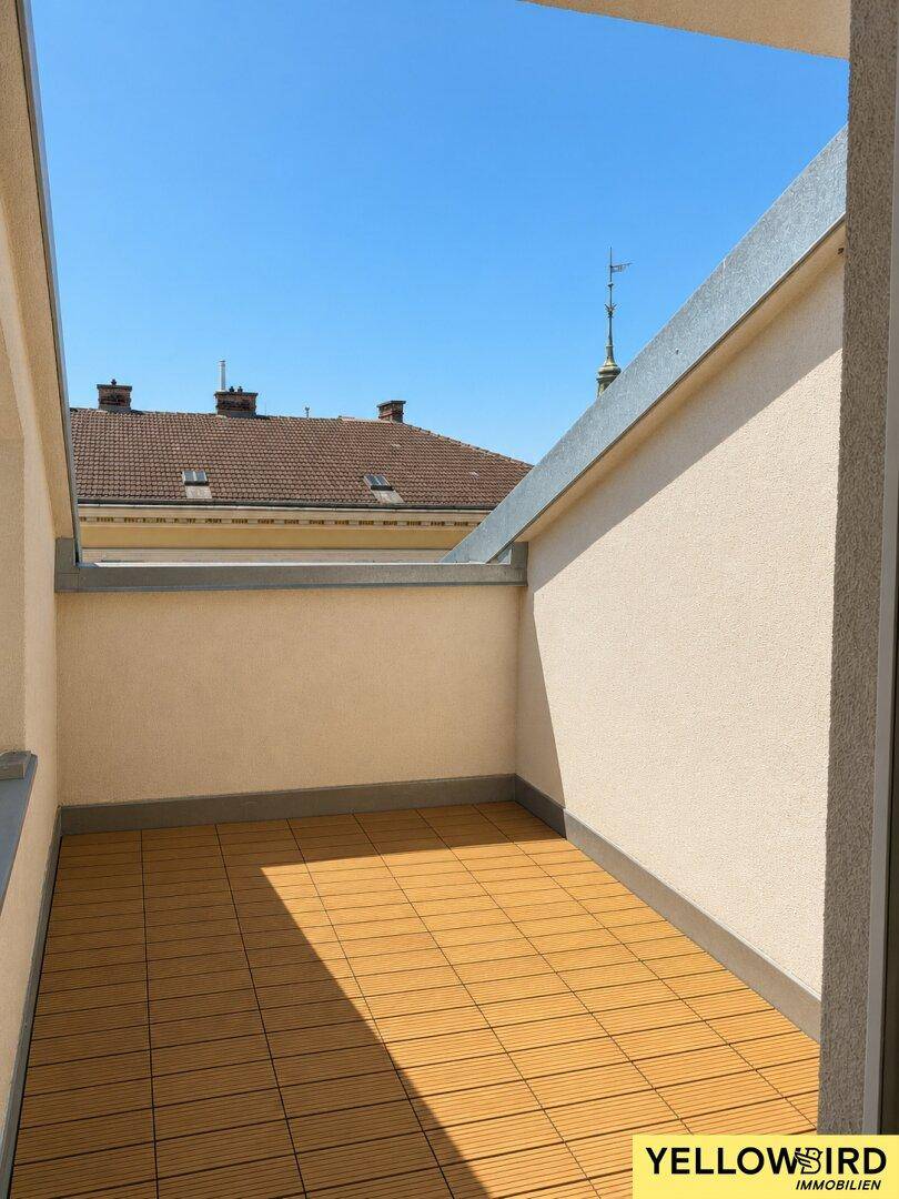 Kleiner, sonniger Balkon mit Holzdielenoptik und Blick auf die Dächer der Stadt.