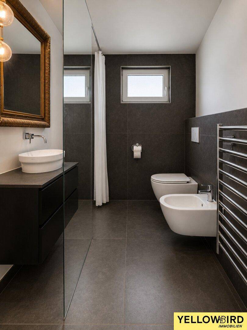 Modernes Badezimmer mit dunklen Fliesen, Bidet, WC und beheiztem Handtuchhalter.