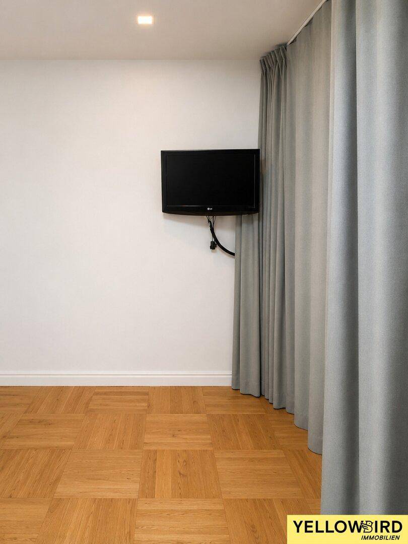 Heller Raum mit Parkettboden und einem an der Wand montierten Fernseher.