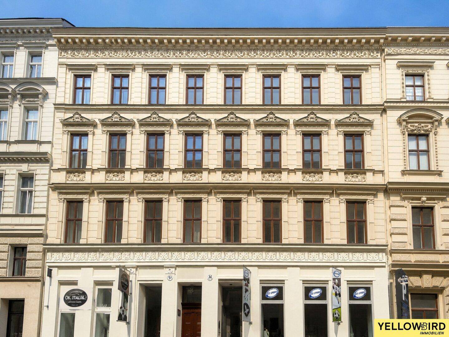 Klassische Fassade eines Altbaus mit vielen Fenstern und dekorativen Elementen unter blauem Himmel.