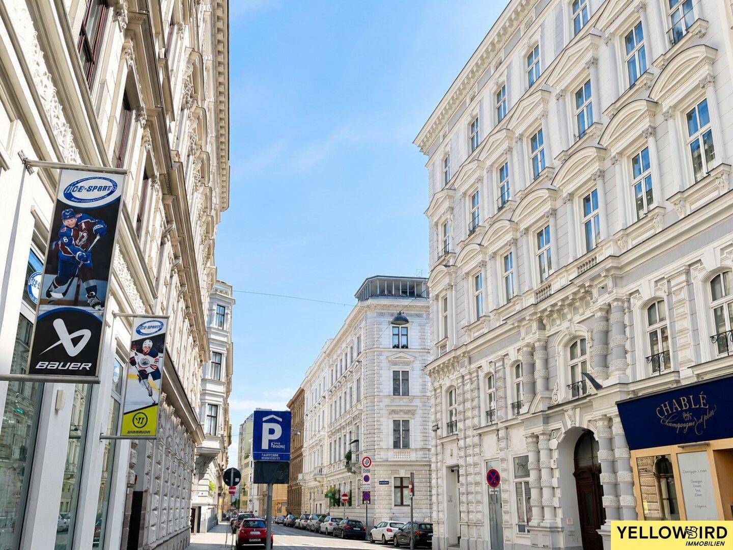 Lebhafte Stadtstraße mit historischen Gebäuden, Geschäften und geparkten Autos unter blauem Himmel.