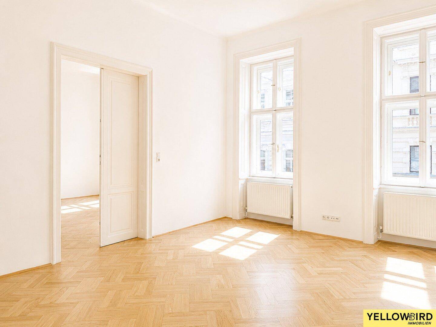Geräumiger, heller Raum mit Fischgrätparkett und zwei großen Fenstern, die viel Tageslicht hereinlassen.