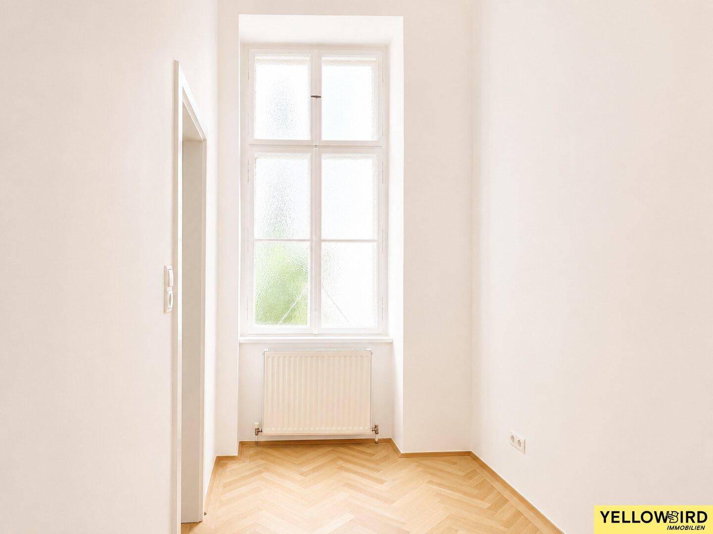 Schmaler, heller Raum mit Fischgrätparkett und einem hohen Fenster, das den Blick ins Grüne freigibt.