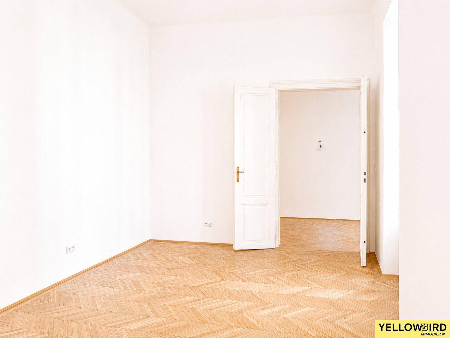 Leerer Raum mit Fischgrätparkett und einer offenen Tür, die den Blick in einen weiteren Raum ermöglicht.
