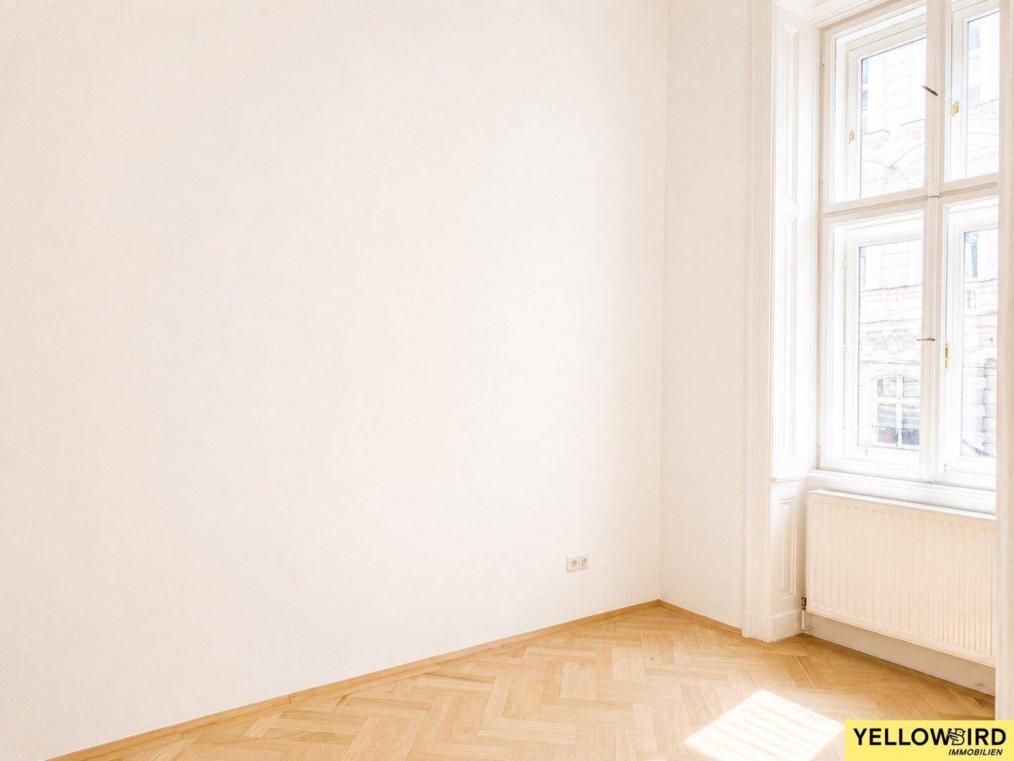 Heller Raum mit Fischgrätparkett und einem hohen Fenster, das viel natürliches Licht in den Raum bringt.
