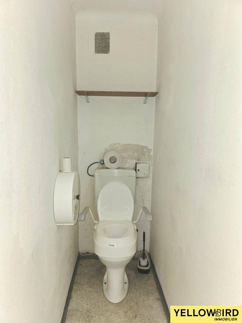 Kleines WC mit Toilette, Ablage und Lüftungsgitter, ausgestattet mit Haltegriffen.