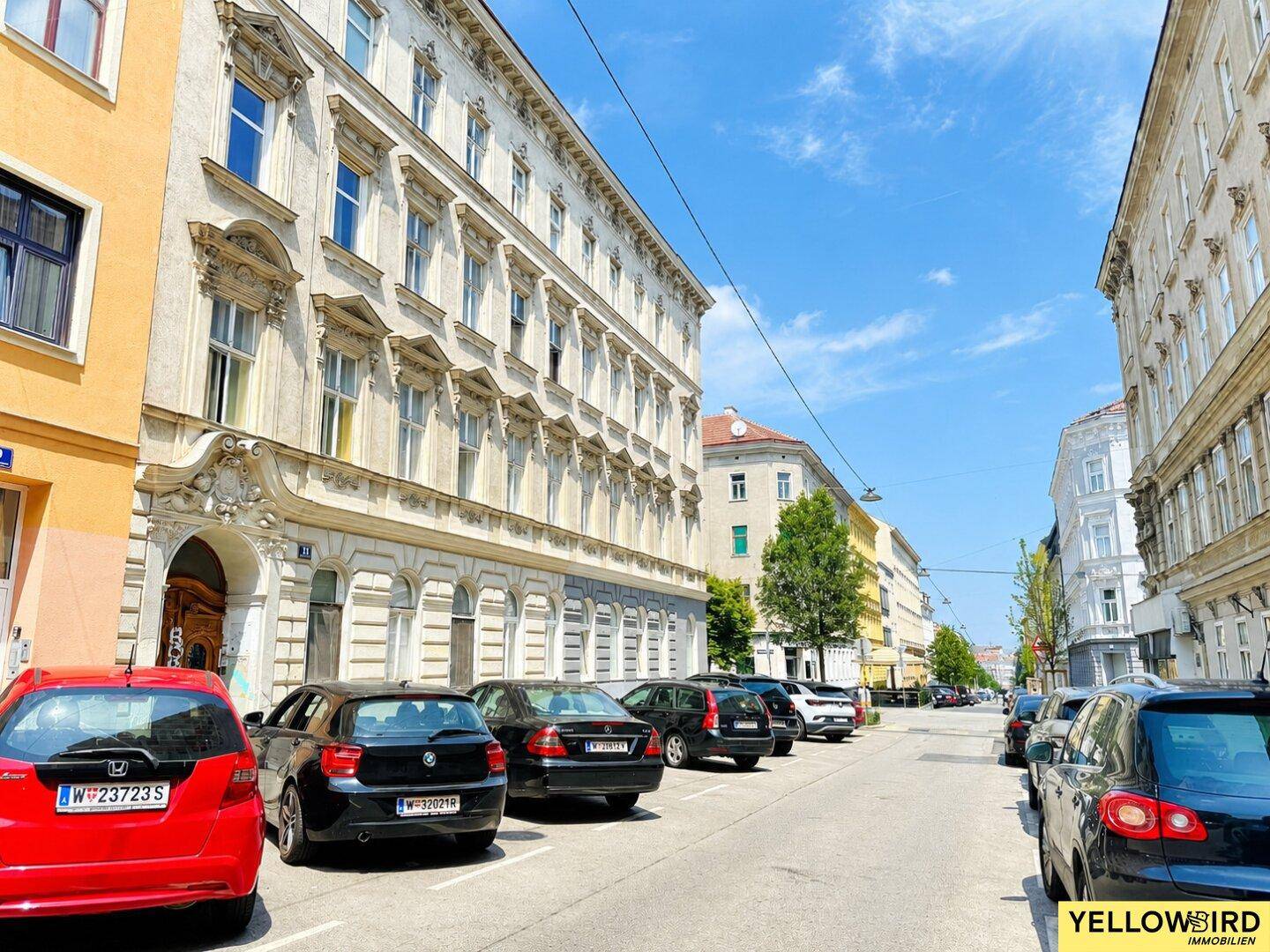 Straße mit parkenden Autos und historischen Gebäuden unter blauem Himmel.