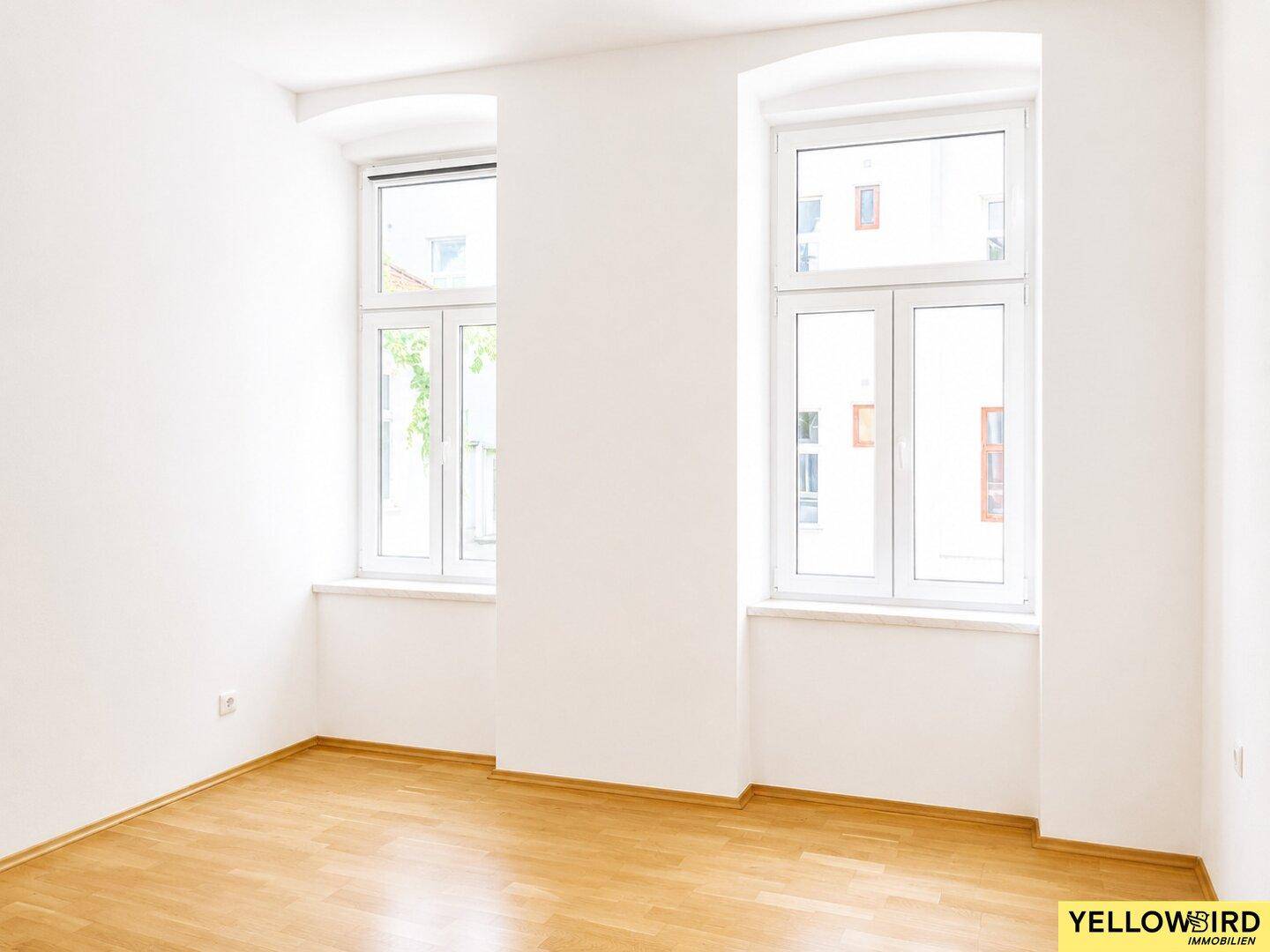 Leerer Wohnraum mit hellem Laminatboden und zwei großen Fenstern.