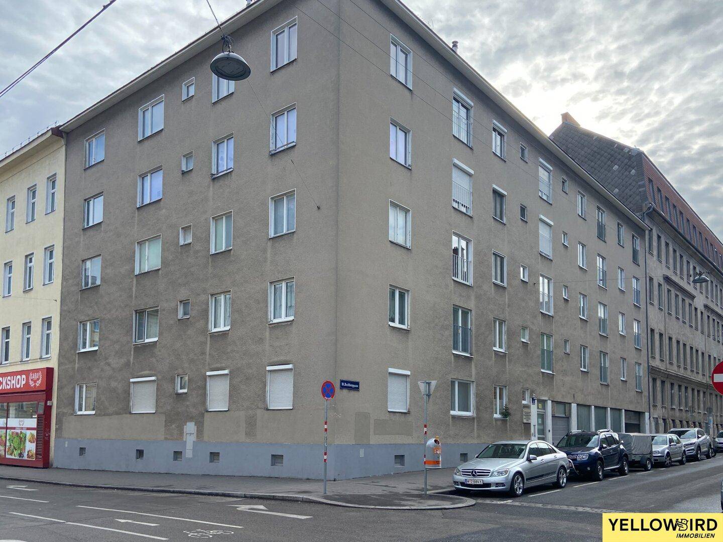 Mehrfamilienhaus in städtischer Umgebung mit zahlreichen Fenstern und parkenden Autos davor.