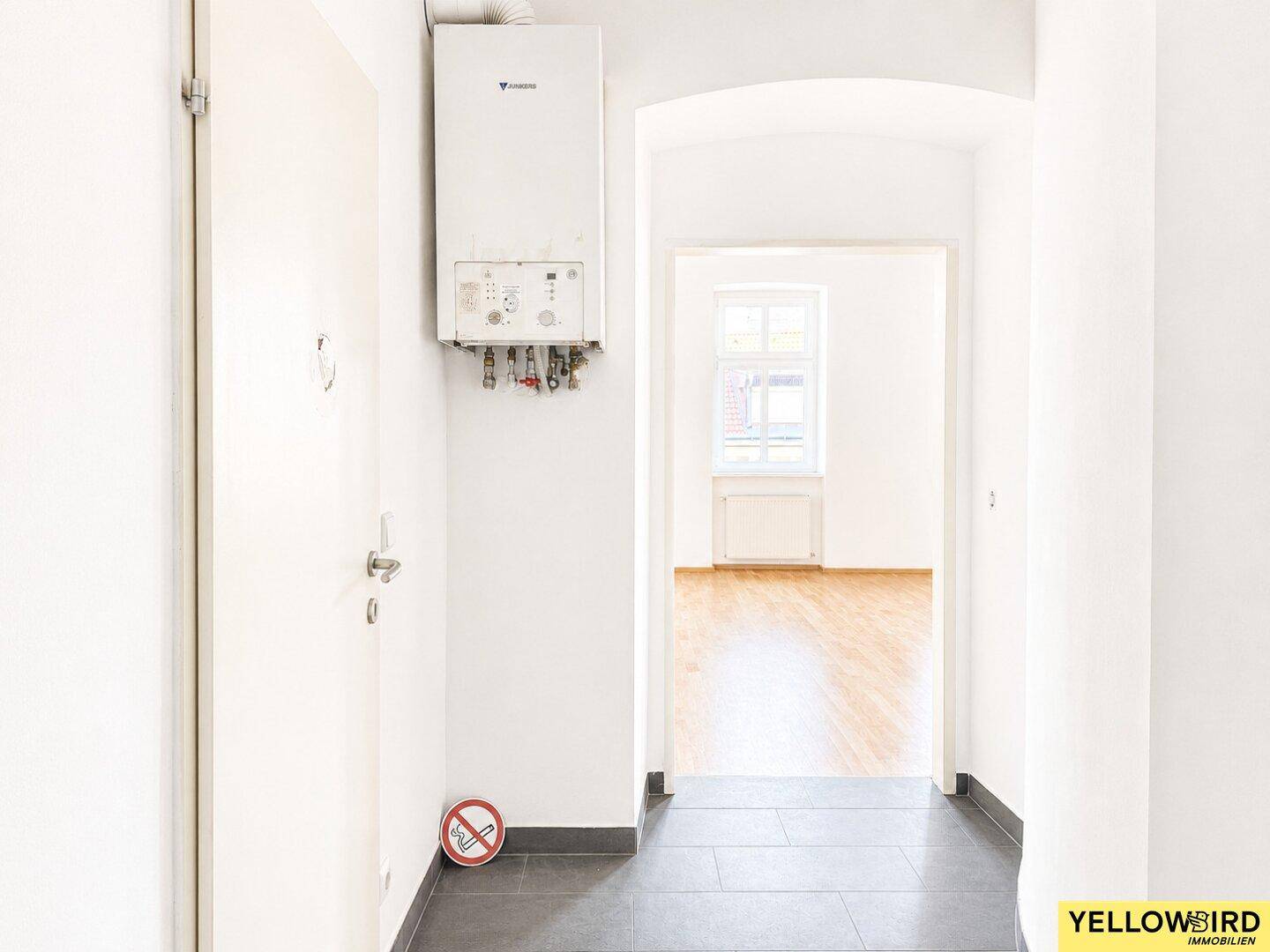 Heller Flur mit Gastherme an der Wand und dunklem Fliesenboden, der in einen weiteren Raum führt.