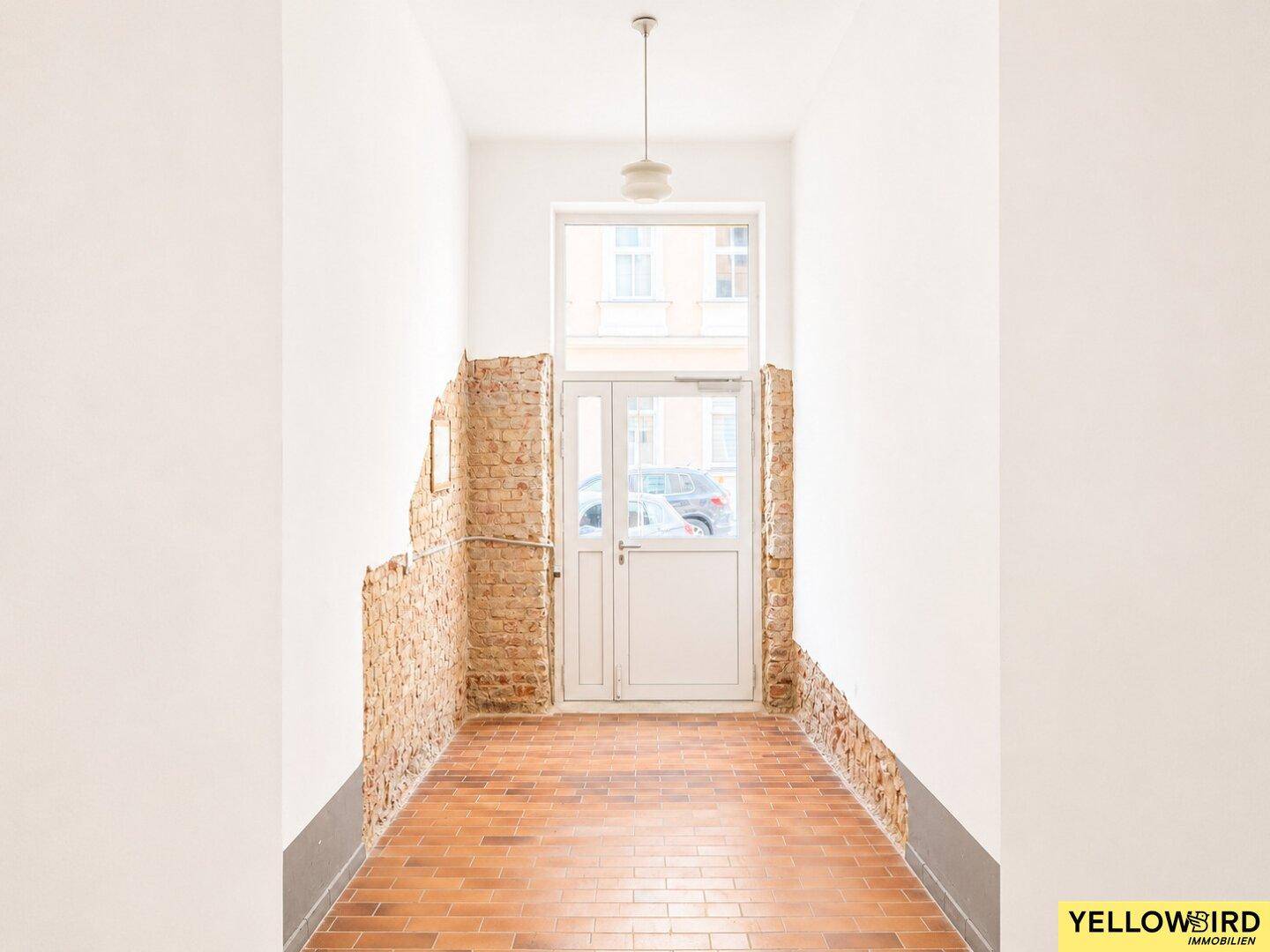 Eingangsbereich mit freigelegten Ziegelwänden, Terrakotta-Bodenfliesen und einer Glastür zur Straße.
