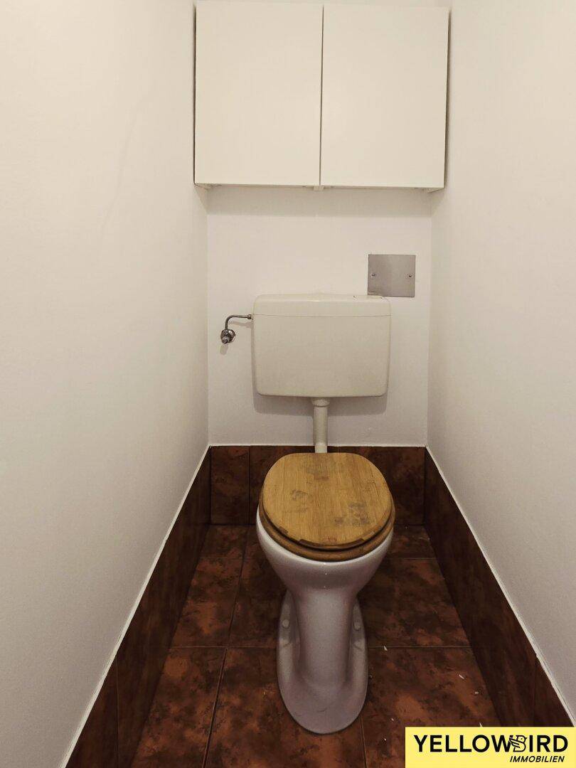 Separates WC mit Toilette, weißen Wänden und braunen Bodenfliesen.