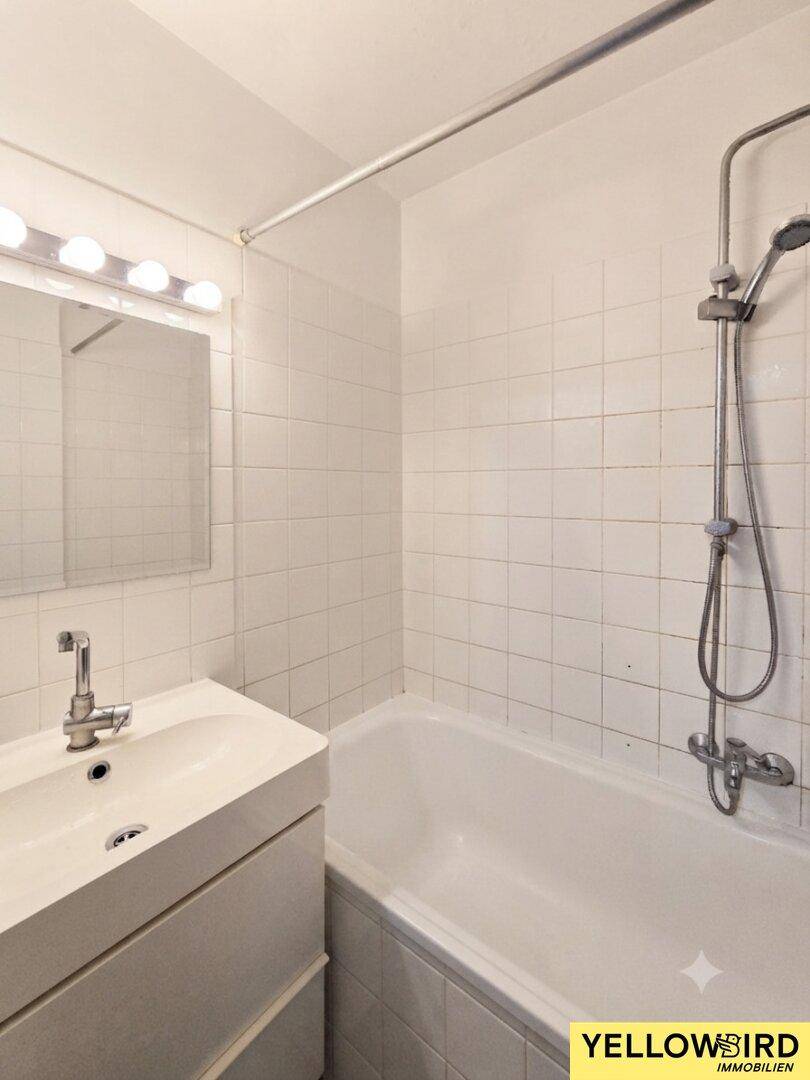 Badezimmer mit weißen Fliesen, Badewanne mit Duschfunktion und Einzelwaschbecken.