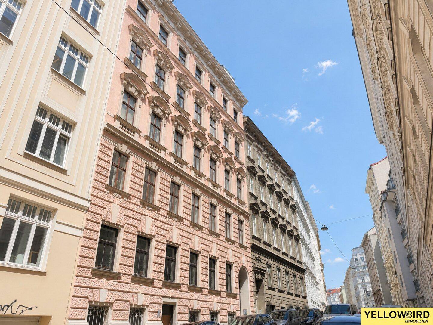 Historische Gebäudezeile mit rosa Fassade unter blauem Himmel in einer belebten Stadtstraße.