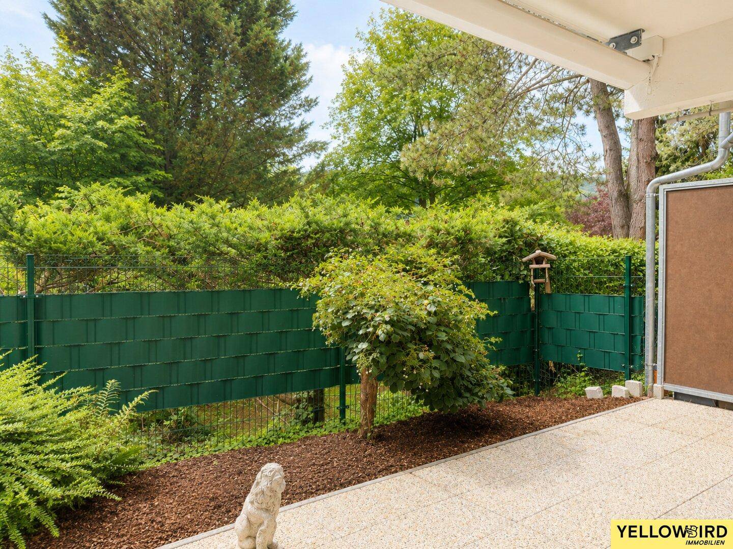 Sonniger Terrassenbereich mit Steinplatten und üppiger grüner Hecke im Hintergrund.
