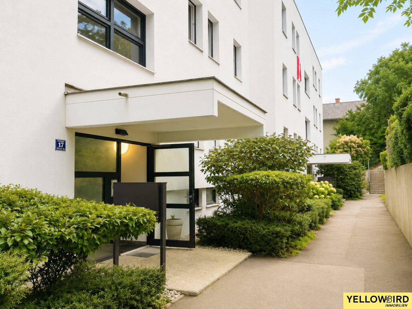 Eingangsbereich eines modernen weißen Mehrfamilienhauses mit gepflegter Bepflanzung.