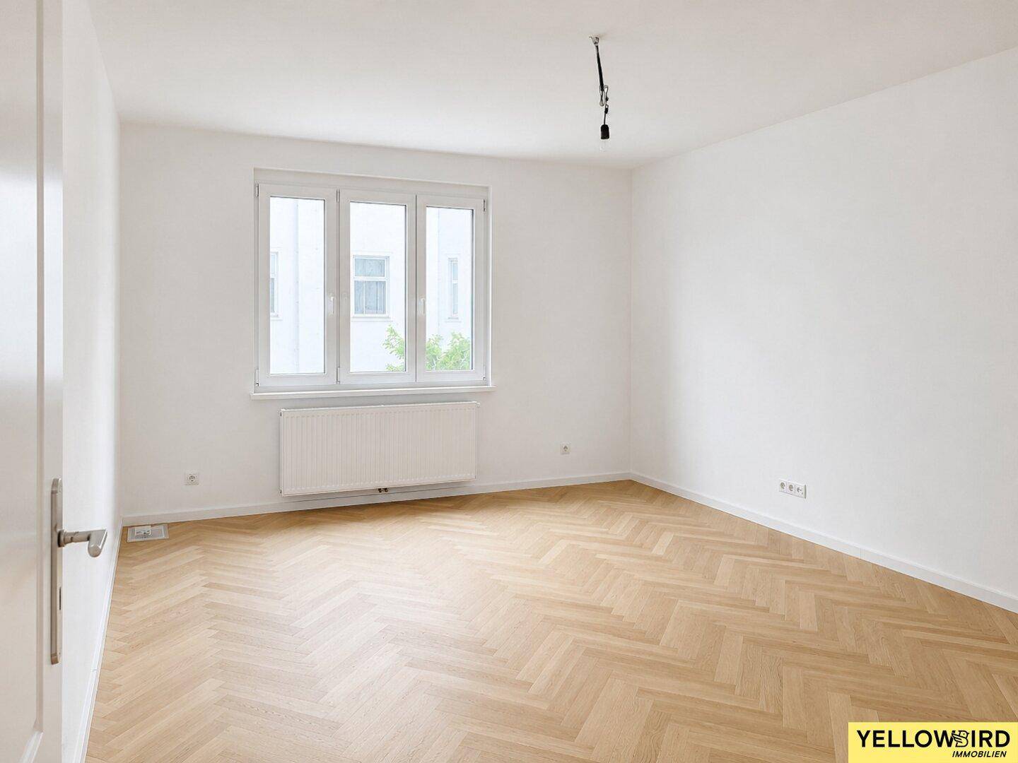 Geräumiges, unmöbliertes Zimmer mit hellem Fischgrätparkett und Fenster, ideal als Schlafzimmer.
