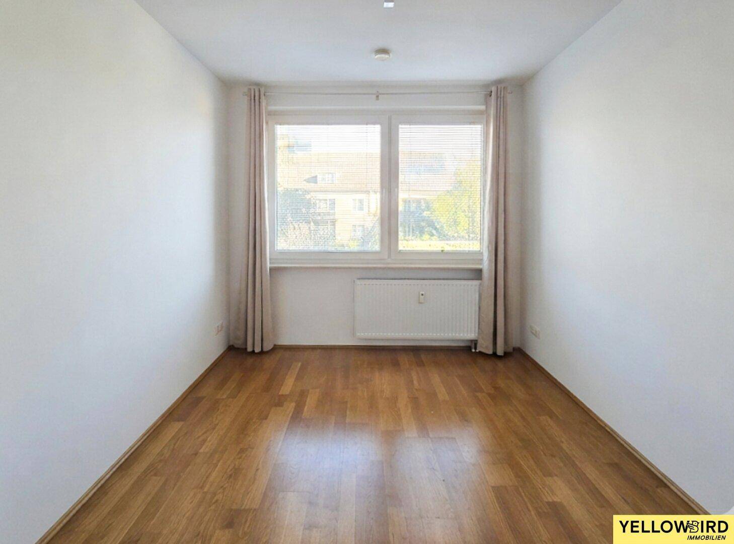 Helles, leeres Zimmer mit Parkettboden und großem Fenster, ideal als Schlaf- oder Arbeitszimmer.