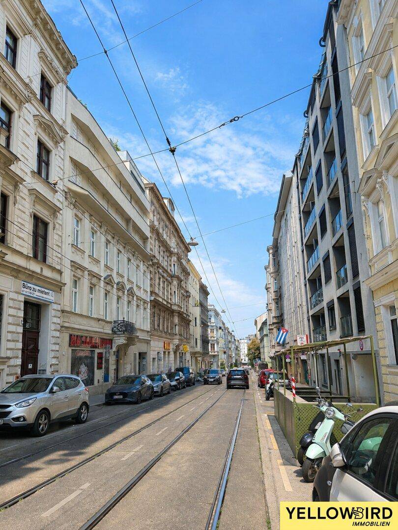 Eine belebte Stadtstraße mit historischen Gebäuden, parkenden Autos und Straßenbahnschienen unter blauem Himmel.