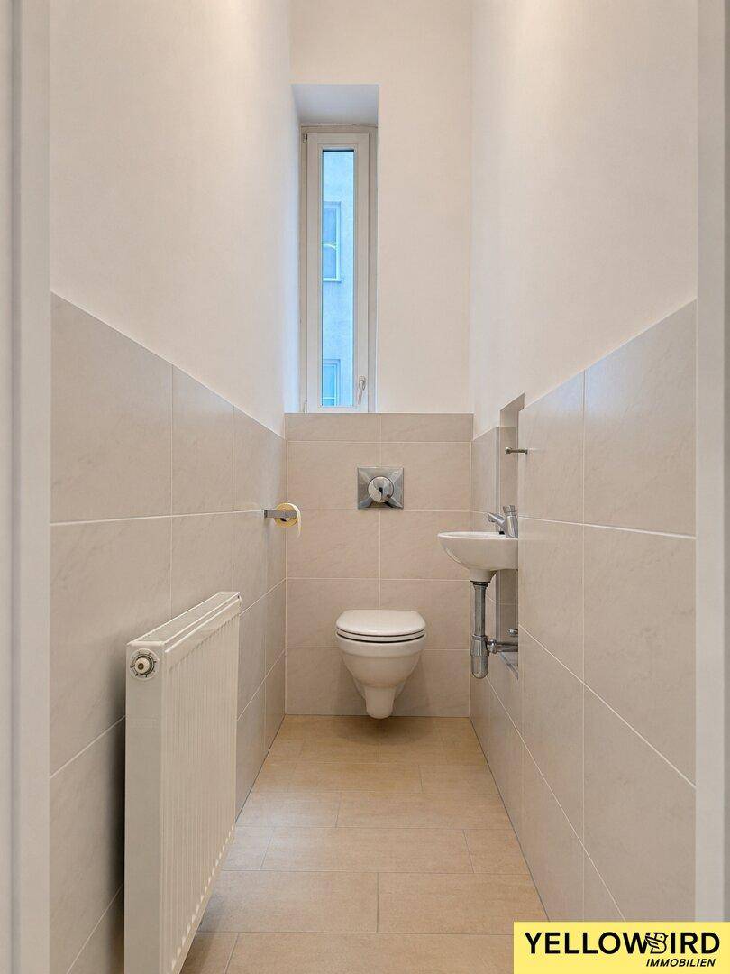 Separates WC mit kleinem Waschbecken, Fenster und Fliesenboden, funktional und hell.