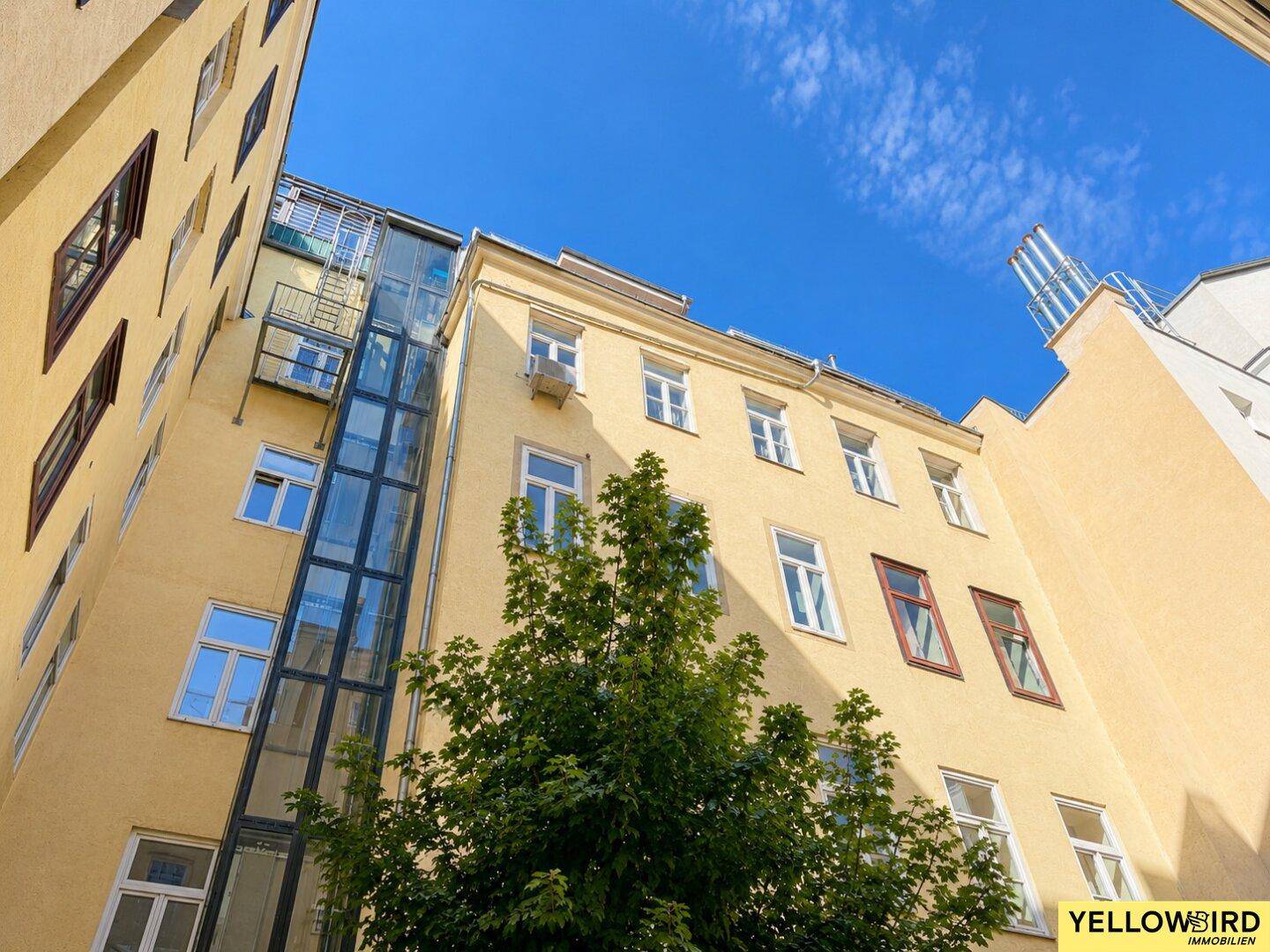 Hinterhofansicht eines Gebäudes mit externem Glasaufzug und grünen Bäumen.