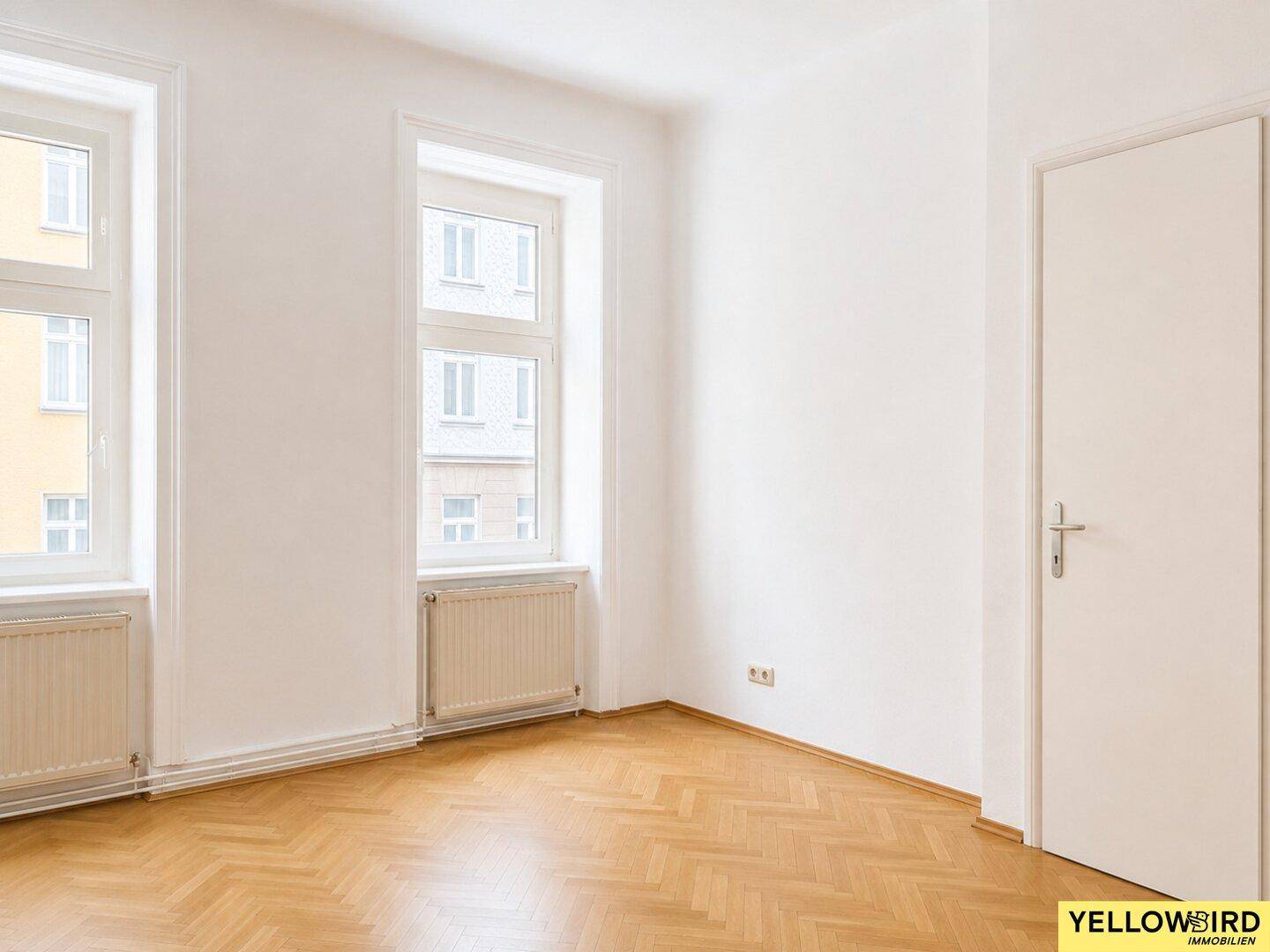 Freundliches Zimmer mit Fischgrätparkett, Heizkörpern unter den Fenstern und weißer Tür.