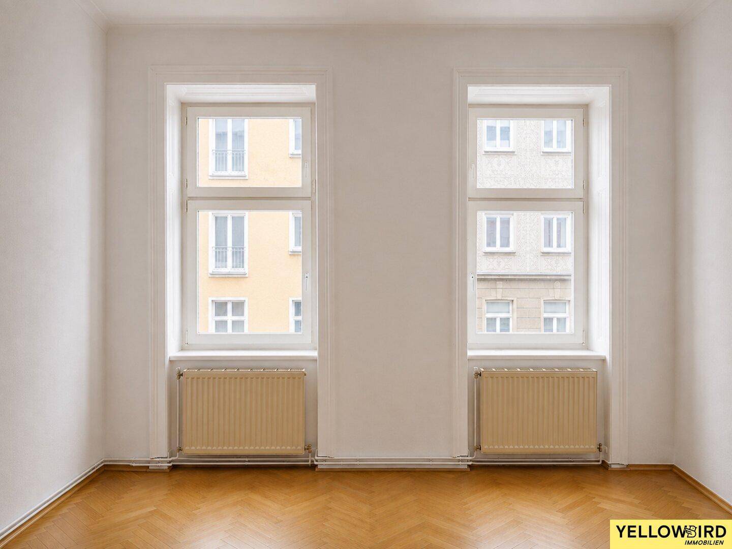 Geräumiges Zimmer mit Fischgrätparkett und zwei großen Fenstern, die viel Licht hereinlassen.