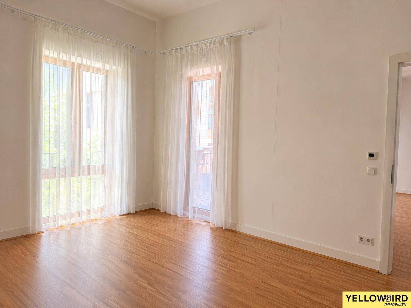 Helles Zimmer mit Holzboden und großen Fenstern, ausgestattet mit leichten Vorhängen für viel Tageslicht.