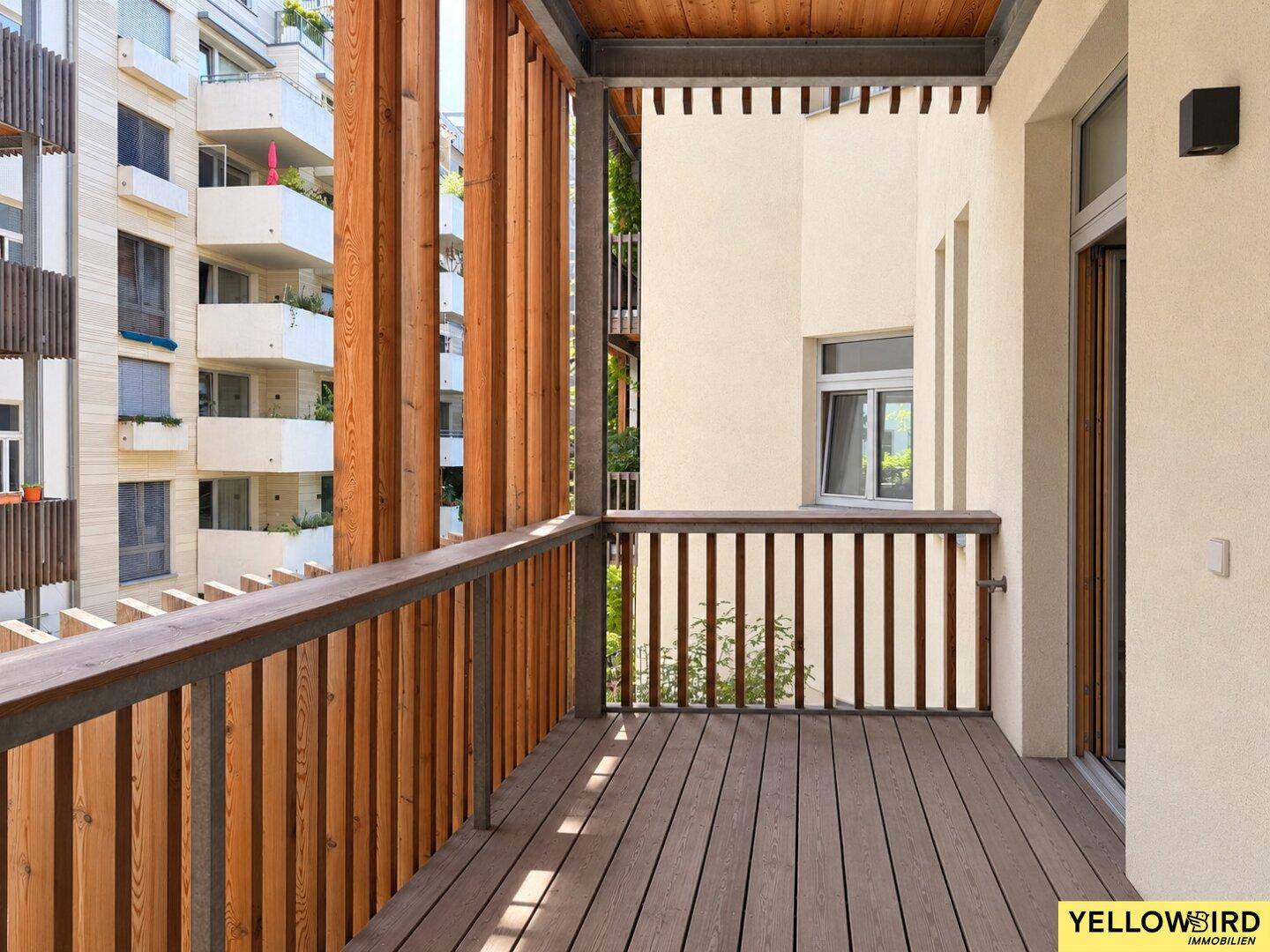 Geräumiger Balkon mit Holzboden und vertikalen Holzlamellen, bietet einen Blick auf umliegende Gebäude.