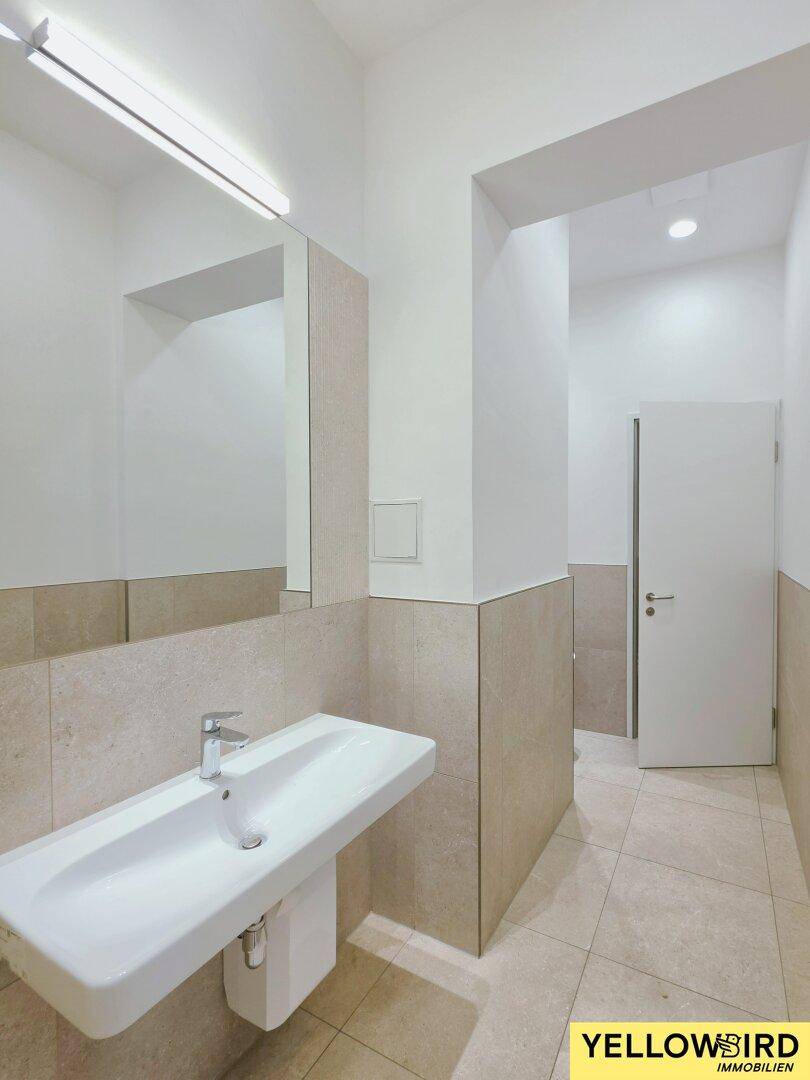 Modernes Badezimmer mit hellem Fliesenboden, Waschbecken und großem Spiegel, das eine saubere Ästhetik bietet.