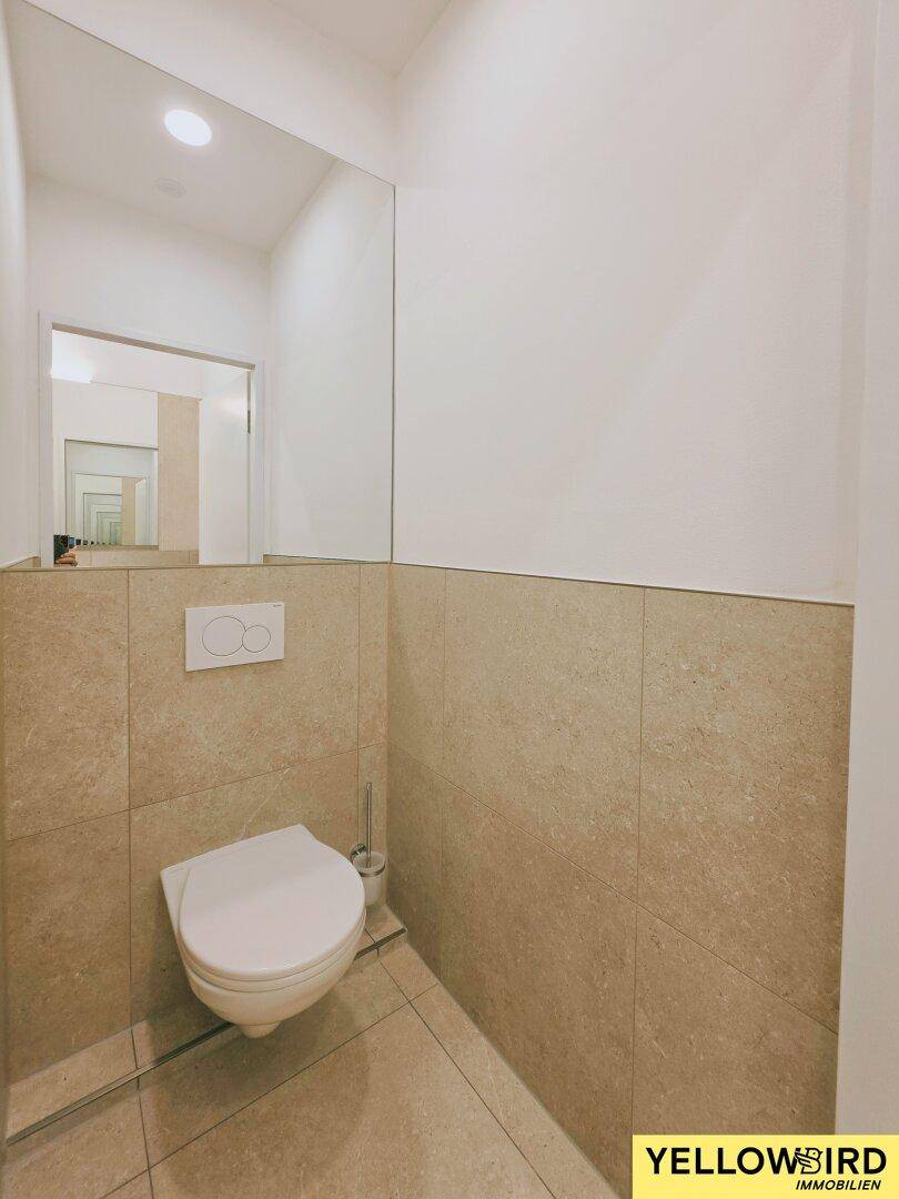 Modernes WC mit Wand-WC, hellem Fliesenboden und großem Spiegel, das eine zeitgemäße Ausstattung aufweist.
