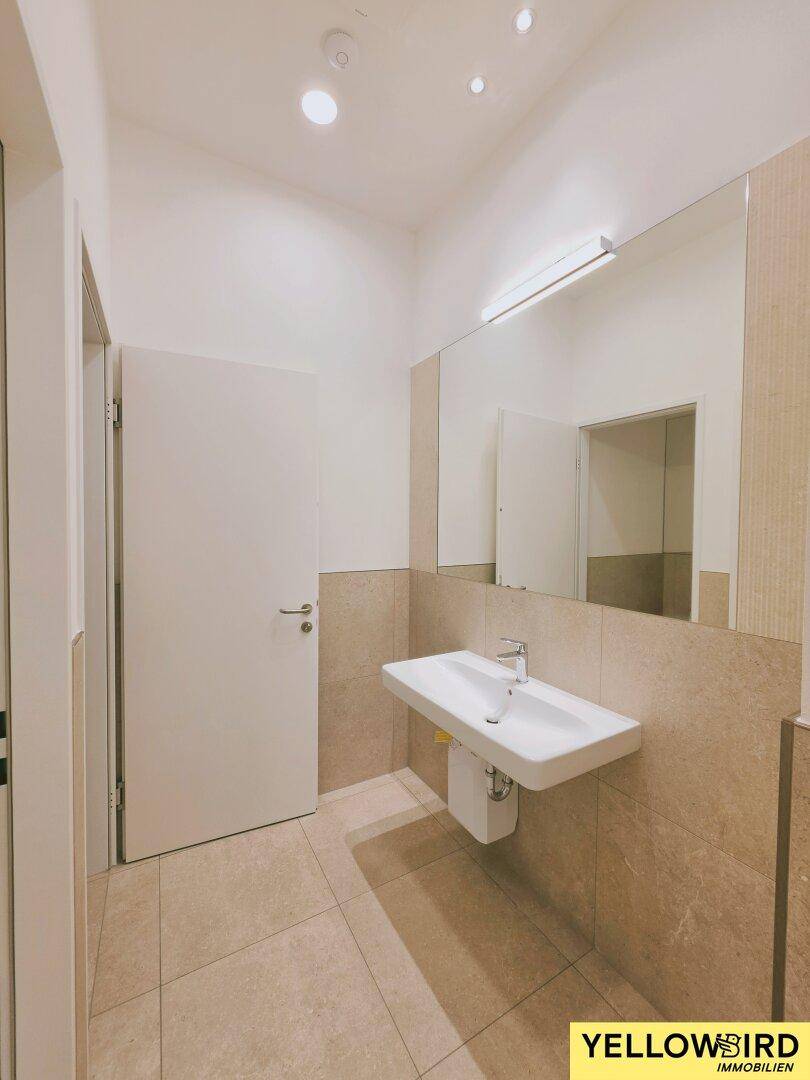 Modernes Badezimmer mit hellem Fliesenboden, Waschbecken und großem Spiegel, das eine funktionale Eleganz bietet.