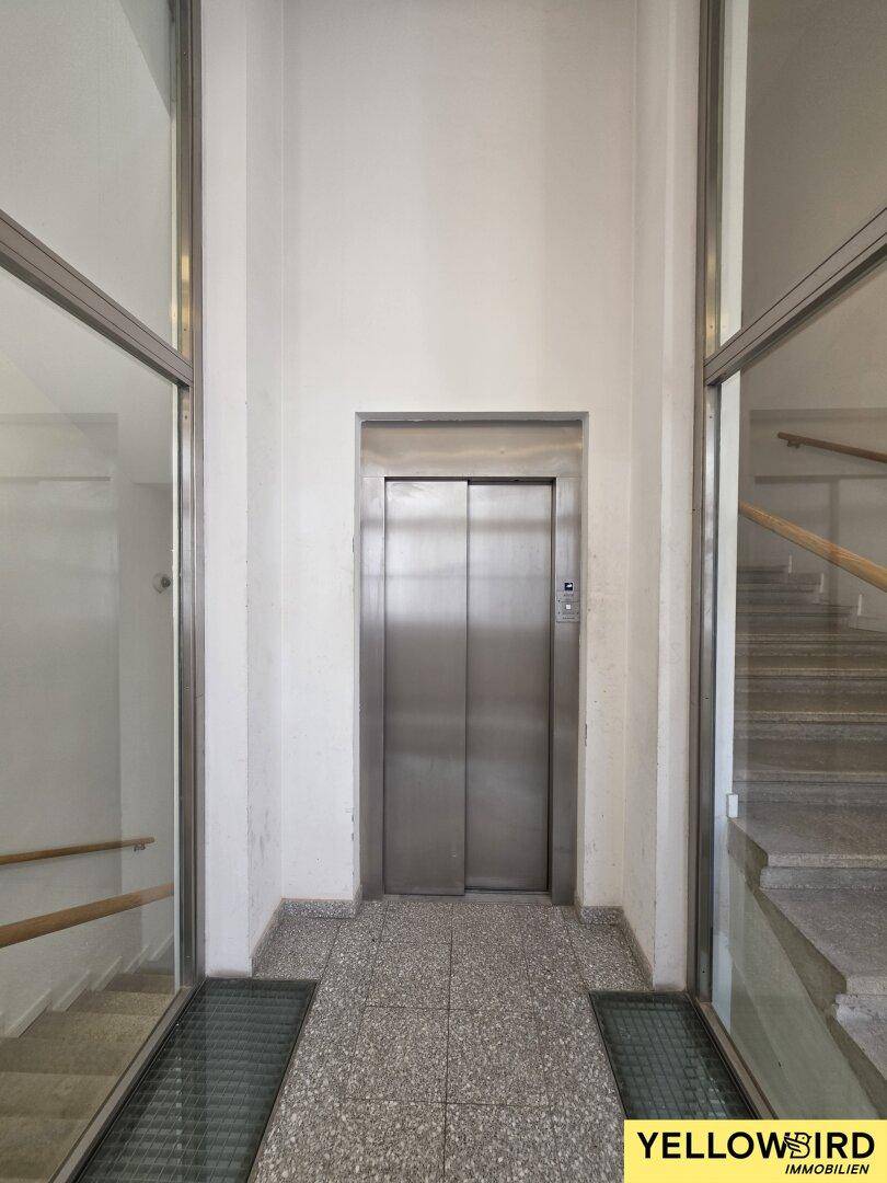 Moderner Aufzug im Treppenhaus mit Edelstahltüren und Glasumrandung, der eine schnelle Verbindung zwischen den Etagen ermöglicht.